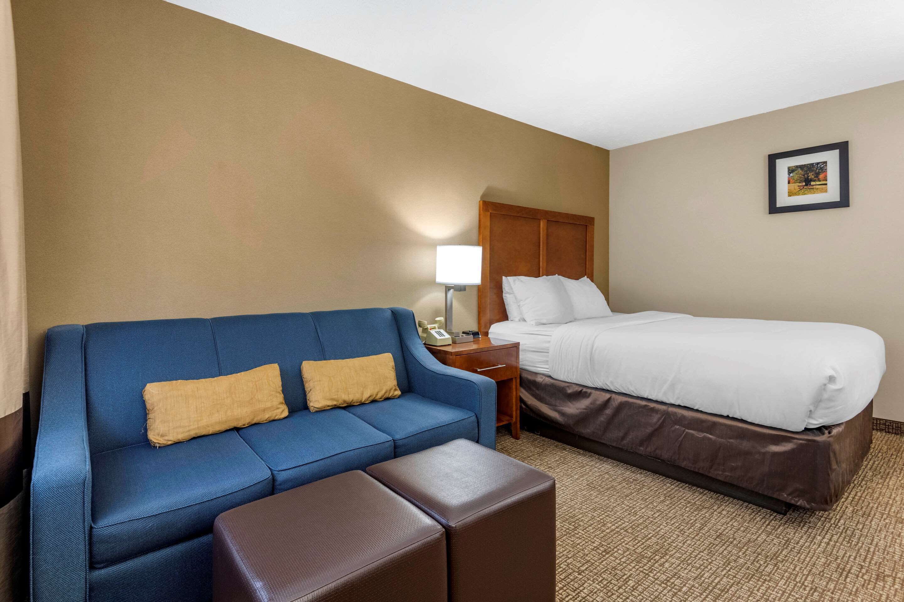 A hotel room with a blue sofa, a brown ottoman, a bed with white bedding, a nightstand with a lamp, and a framed picture on the wall.