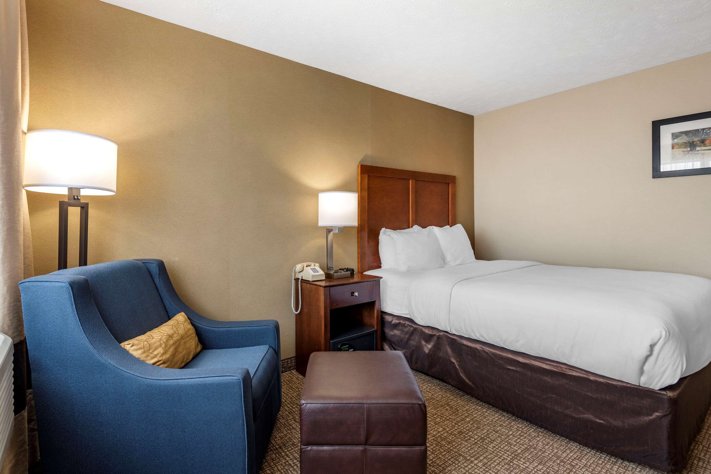Una habitación de hotel con una cama, un sillón azul, una mesita de noche de madera con una lámpara y un otoman marrón.