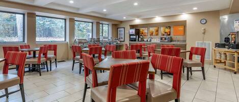 A dining area with tables and chairs, a kitchen area, and large windows with a view of trees.