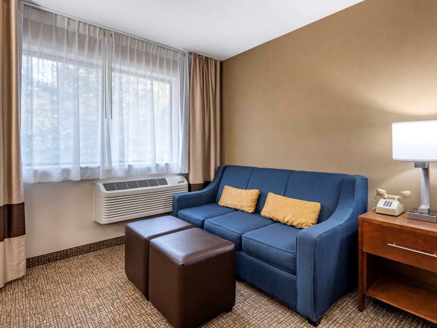 A hotel room with a blue sofa, two brown ottomans, a wooden nightstand with a lamp, a telephone, and an air conditioning unit.