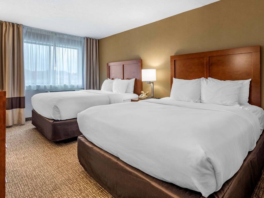 A hotel room with two beds, a wooden headboard, a bedside table with a lamp, and a large window with curtains.