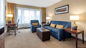 Une chambre d’hôtel avec un canapé bleu, un fauteuil assorti, un bureau avec une lampe et une grande fenêtre avec des rideaux.