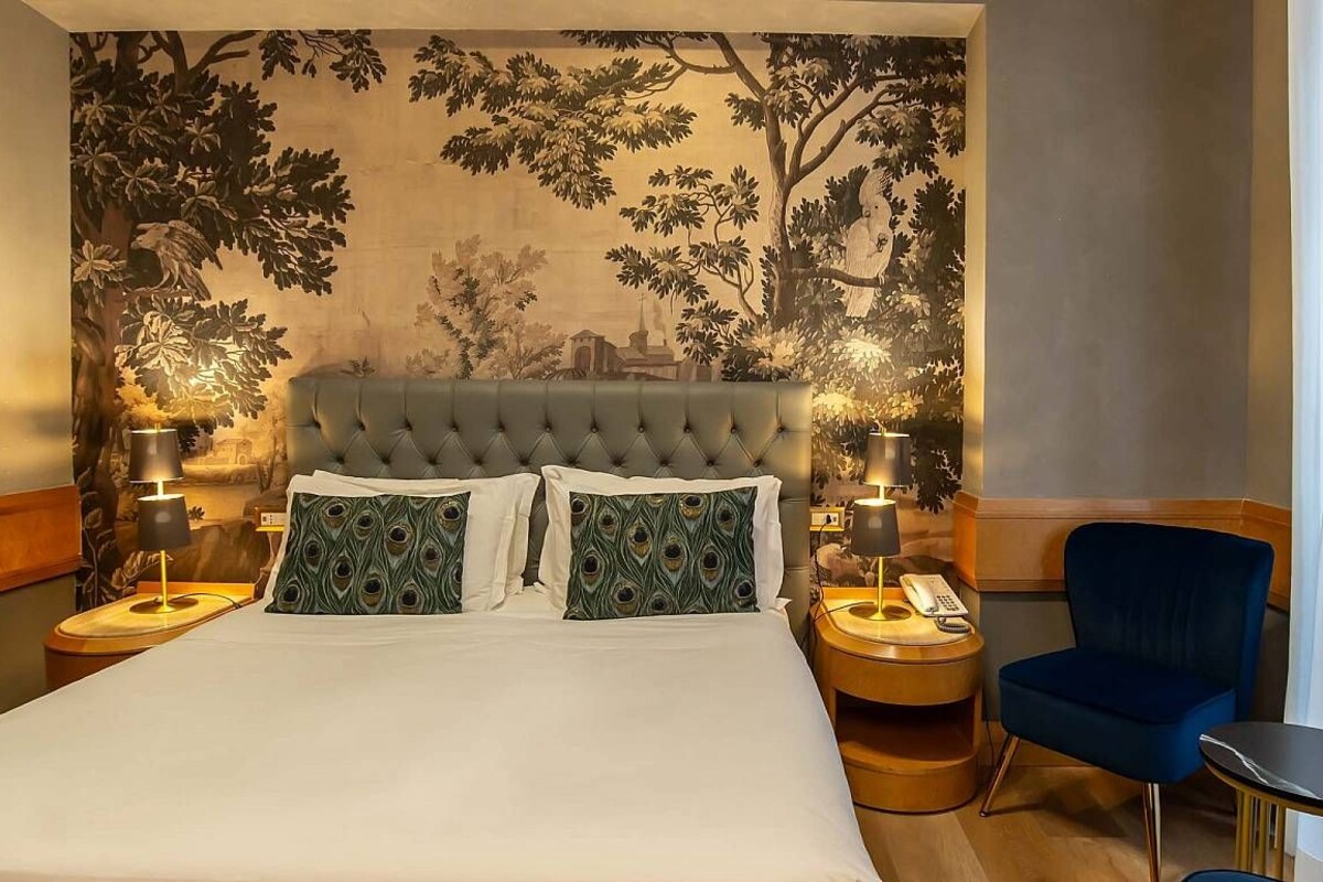 A hotel room with a large bed, two bedside tables, a blue armchair, and a wall mural.