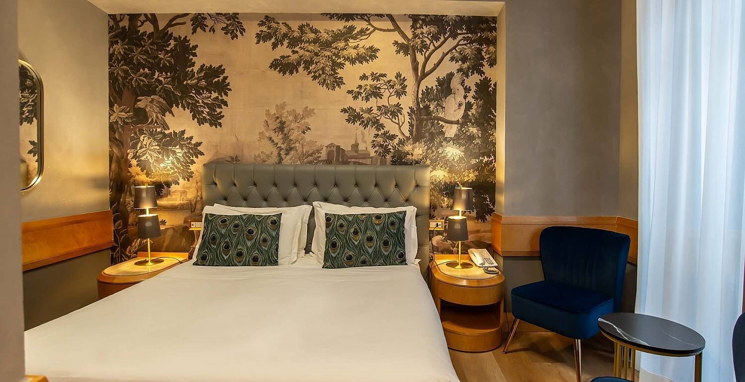 A hotel room with a large bed, two bedside tables, a blue armchair, and a wall mural.