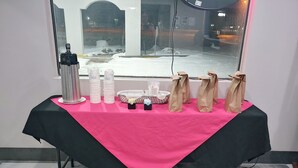 Une table recouverte d’une nappe rose, avec des gobelets en papier blancs, un thermos argenté et des sacs en papier marron.