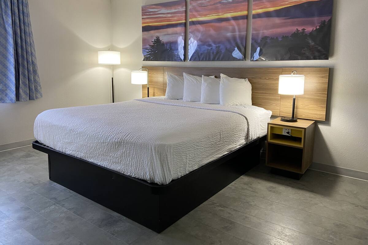 A modern hotel room with a large bed, two bedside tables with lamps, a view of a sunset painting, and a window with curtains.