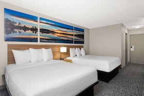 A hotel room with two beds, a wooden headboard, a nightstand with a lamp, and a large landscape painting above the beds.