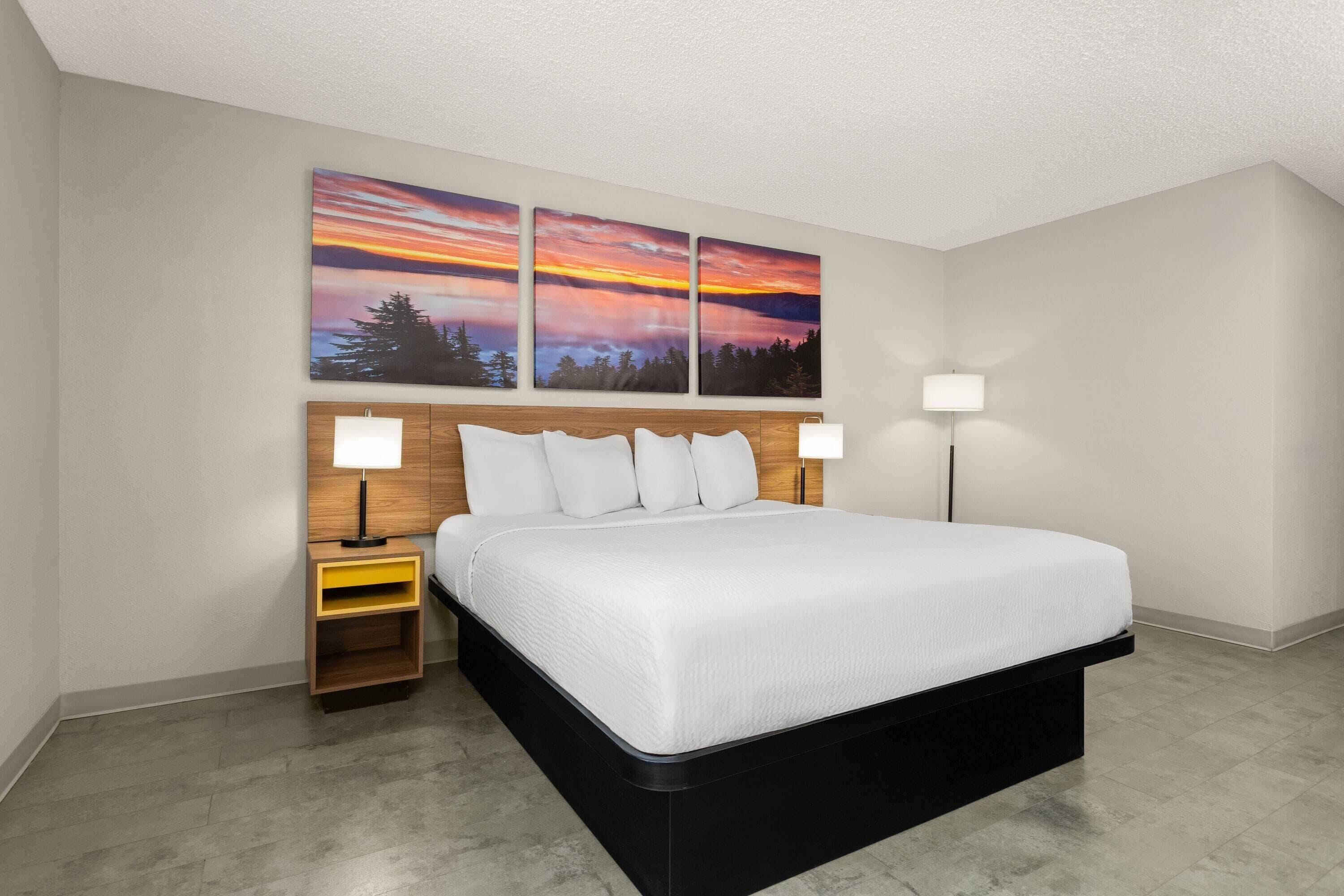 A modern hotel room with a large bed, two bedside tables, and a wall-mounted artwork depicting a sunset.