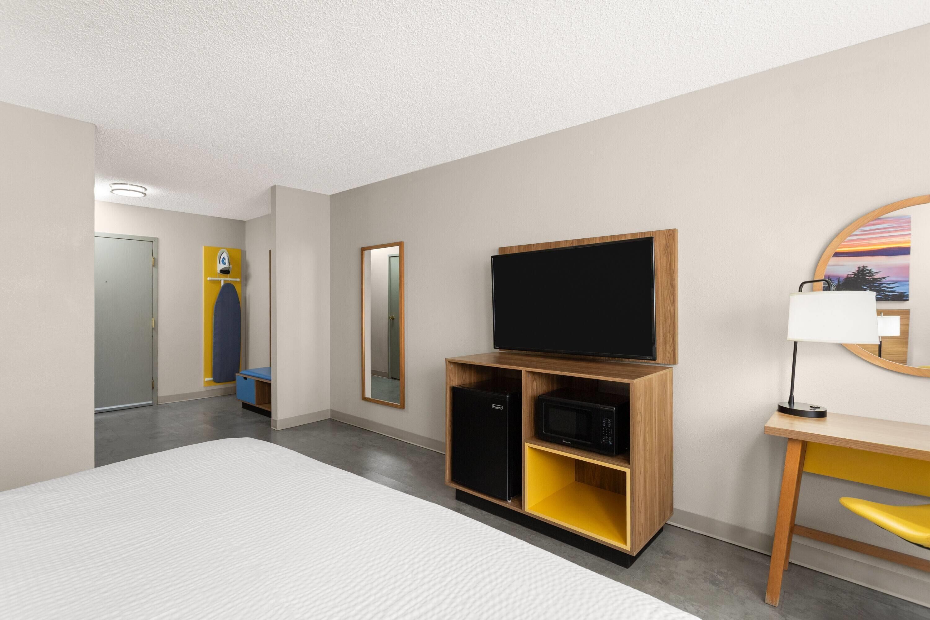 A hotel room with a bed, a TV on a wooden stand, a microwave, a desk with a chair, and a mirror on the wall.