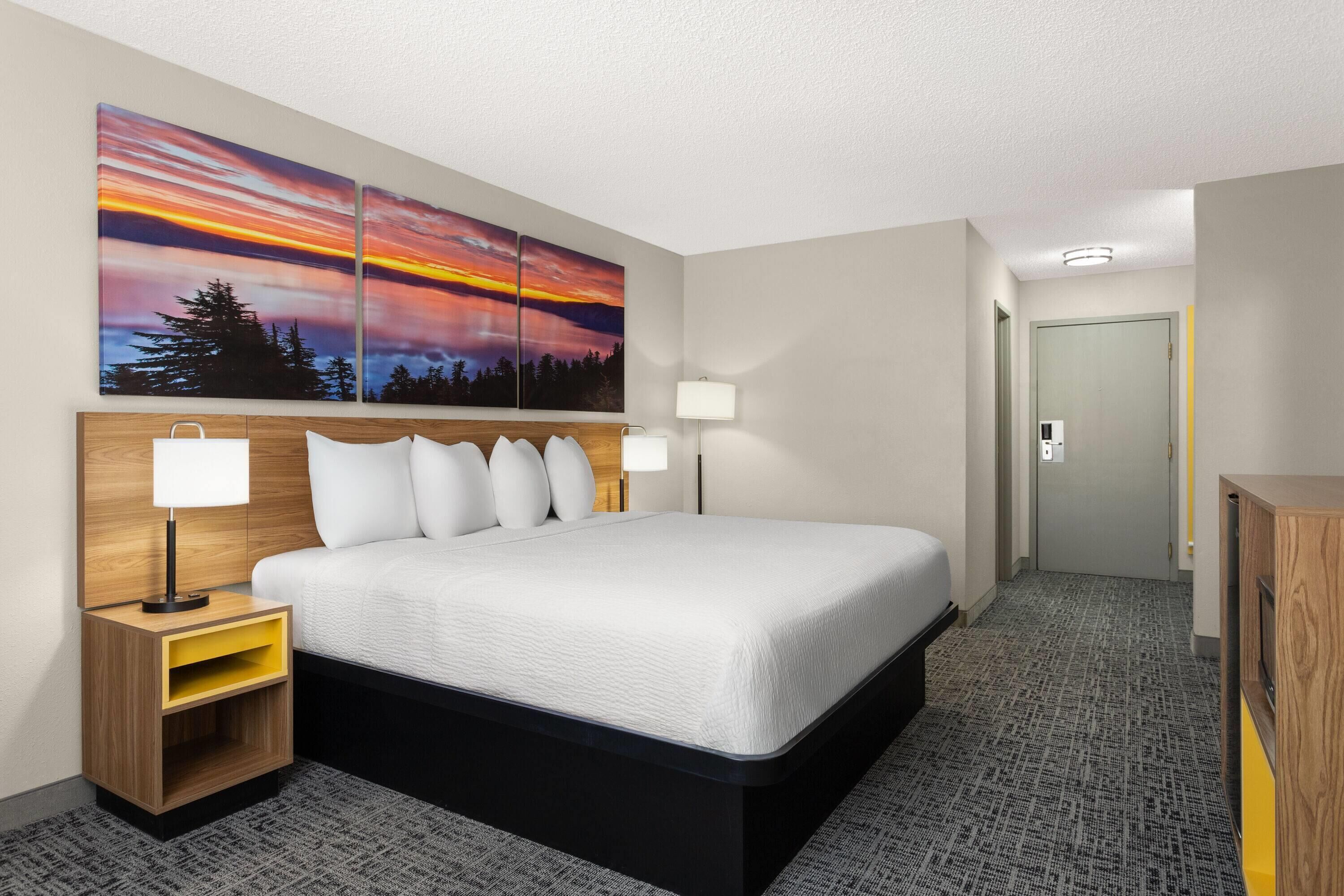 A hotel room with a large bed, two bedside tables with lamps, a nightstand with a storage drawer, and a wall-mounted painting of a sunset.