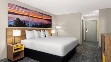 A hotel room with a large bed, two bedside tables with lamps, a nightstand with a storage drawer, and a wall-mounted painting of a sunset.