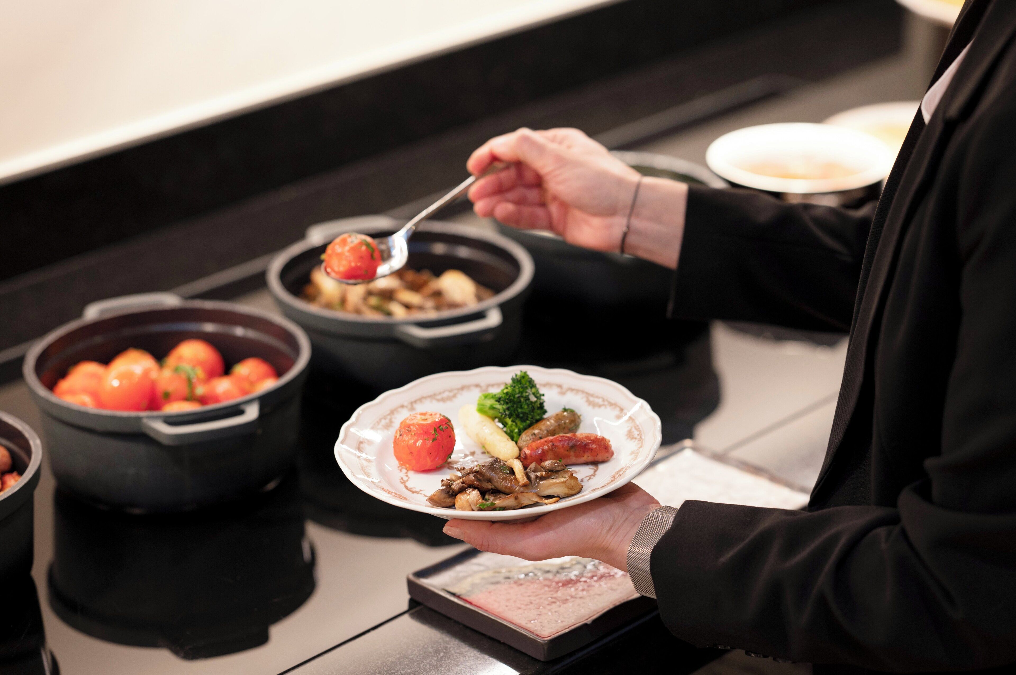A person serving a plate with various food items including a tomato, broccoli, and sausages.