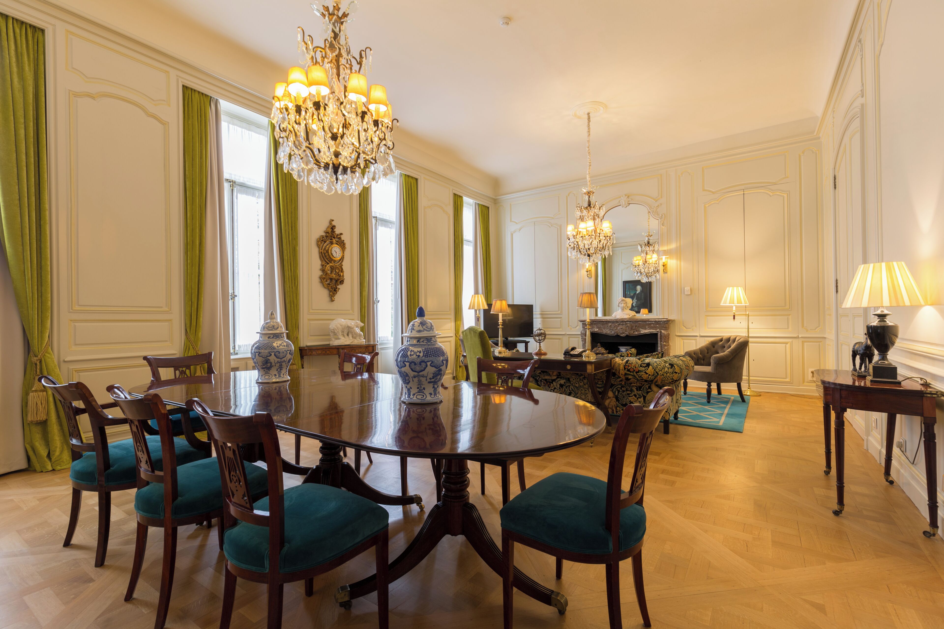 A formal dining room with a round table, chairs, chandeliers, and a television.
