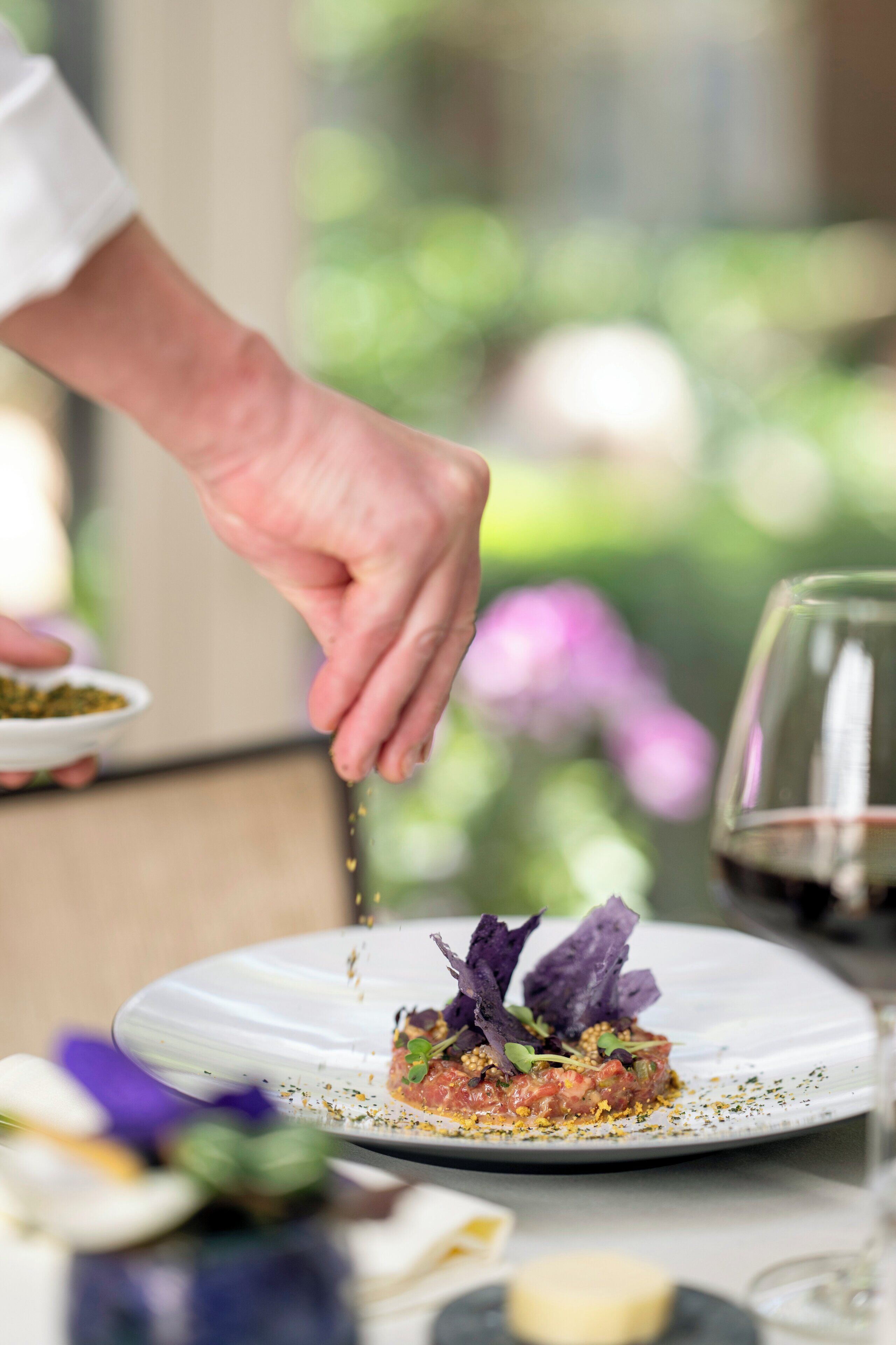 Una mano colocando un plato sobre otro, con un fondo borroso de follaje y flores.