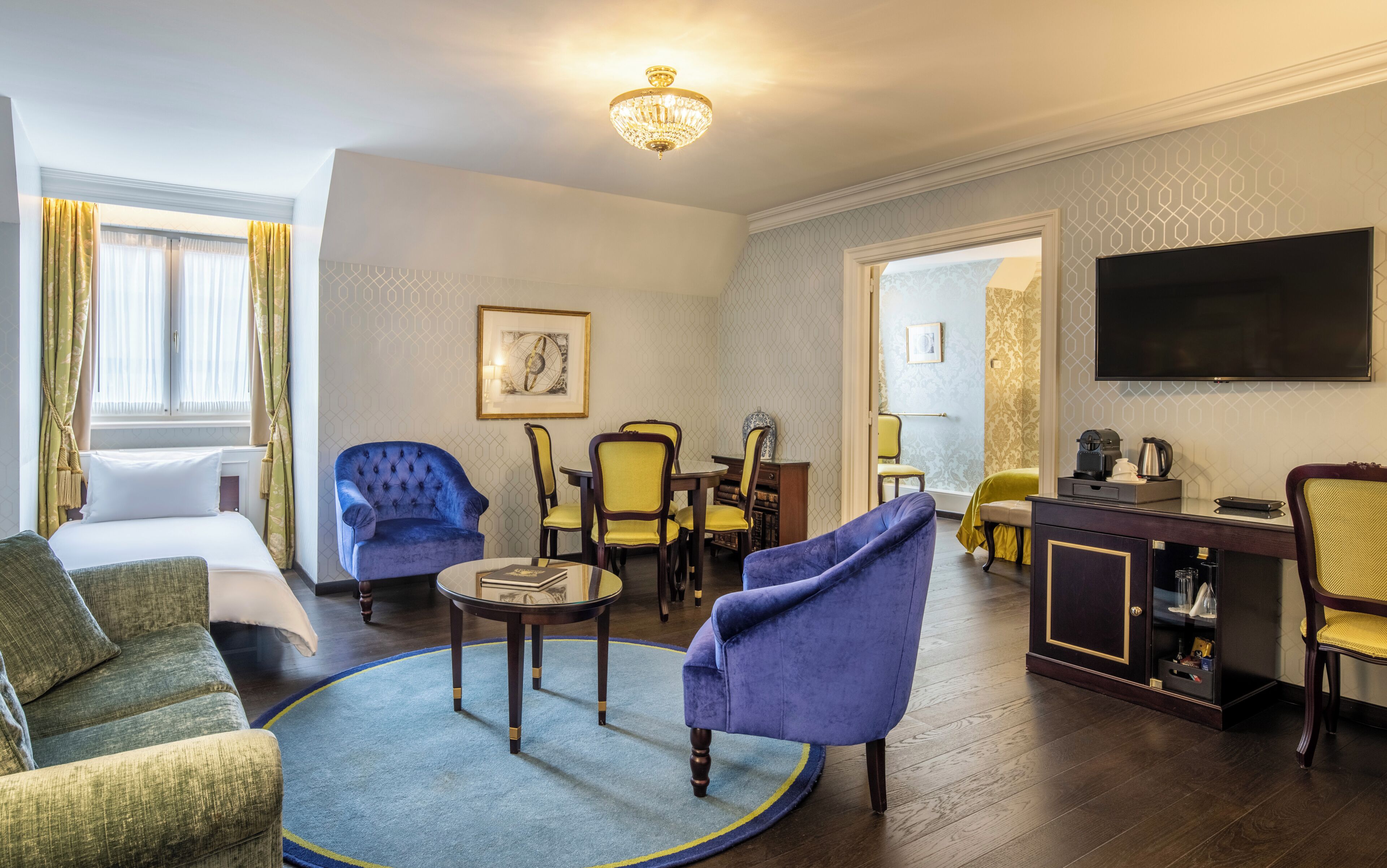 A hotel room with a bed, two armchairs, a round coffee table, a TV, and a desk with a computer.