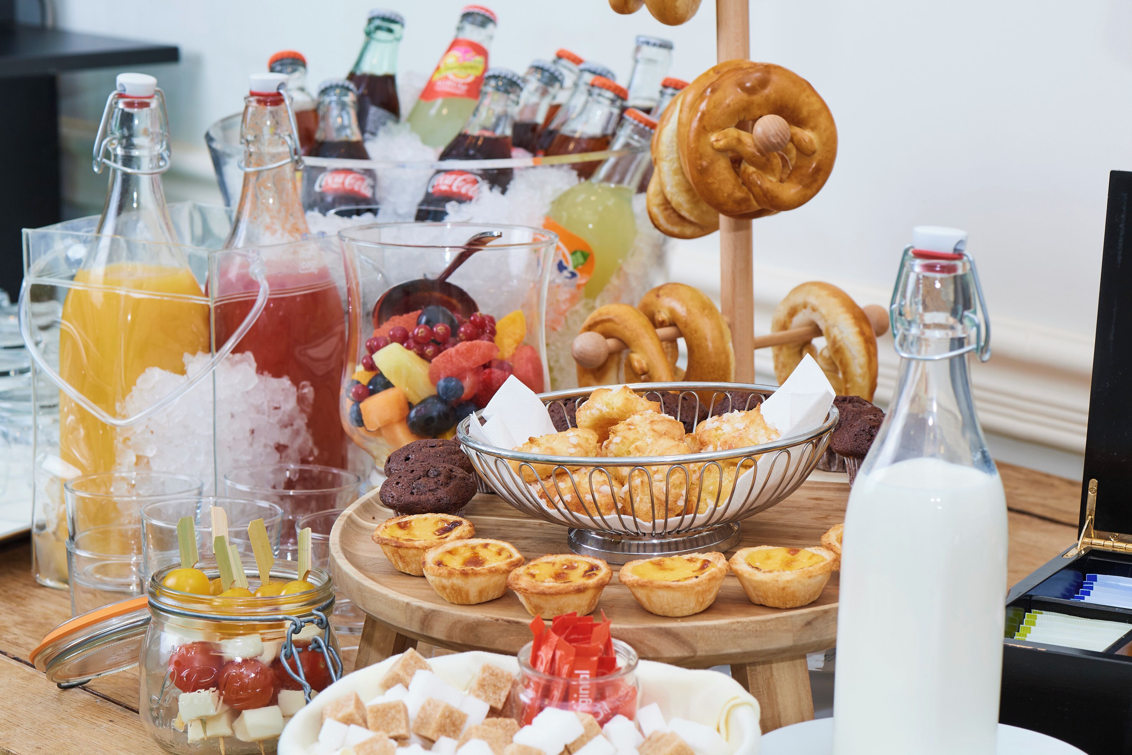 A variety of food items and beverages on a wooden table, including jars of drinks, a bowl of pastries, and a bottle of milk.