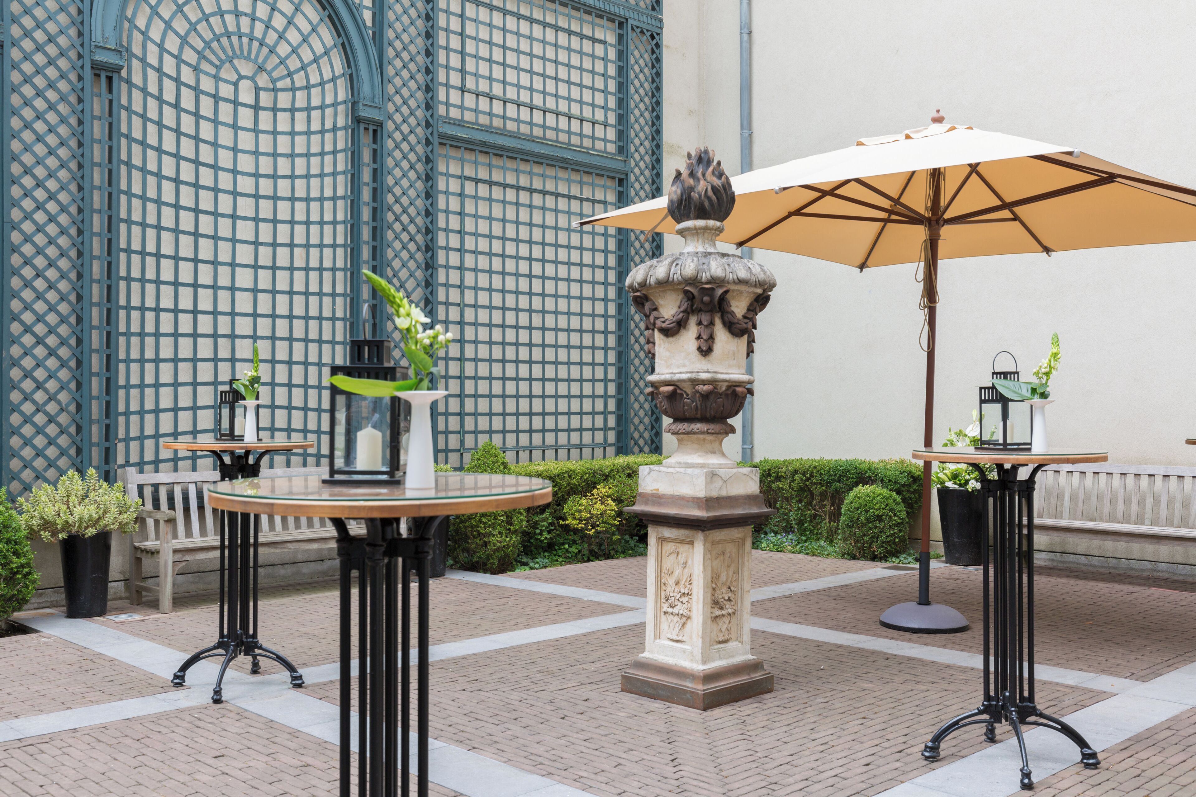 An outdoor patio area with two round tables, one with a beige umbrella, and a decorative stone fountain.