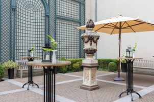 An outdoor patio area with two round tables, one with a beige umbrella, and a decorative stone fountain.