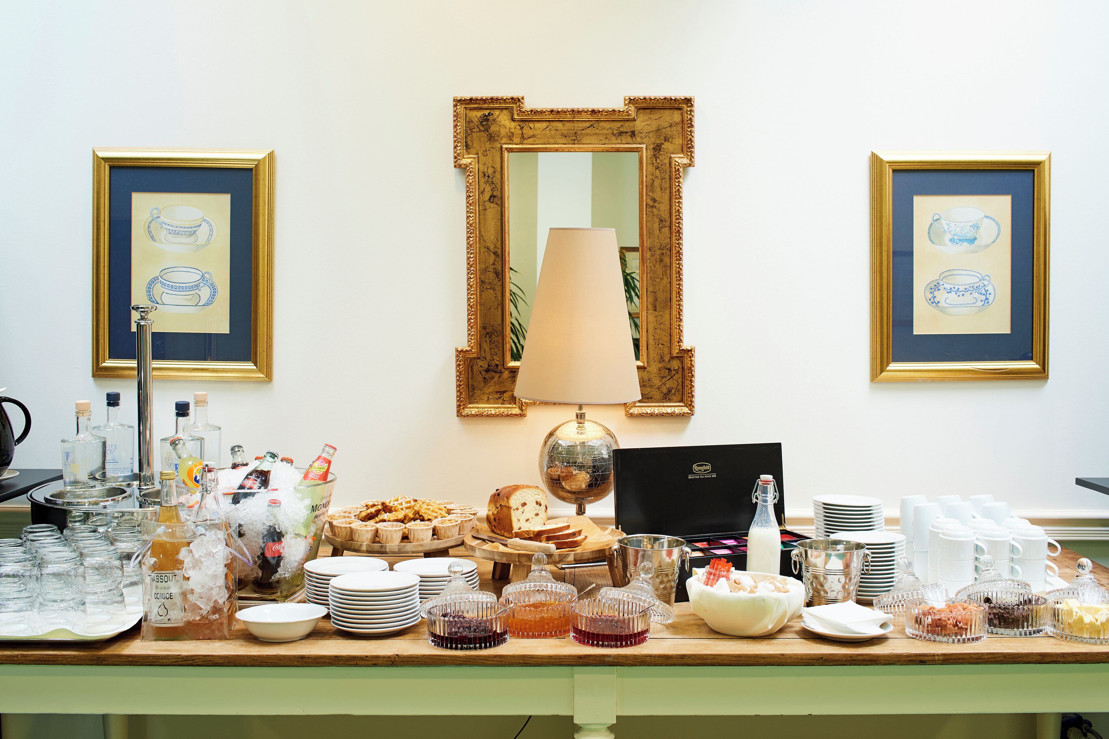 A buffet table with various food items, a lamp, and framed pictures on the wall.