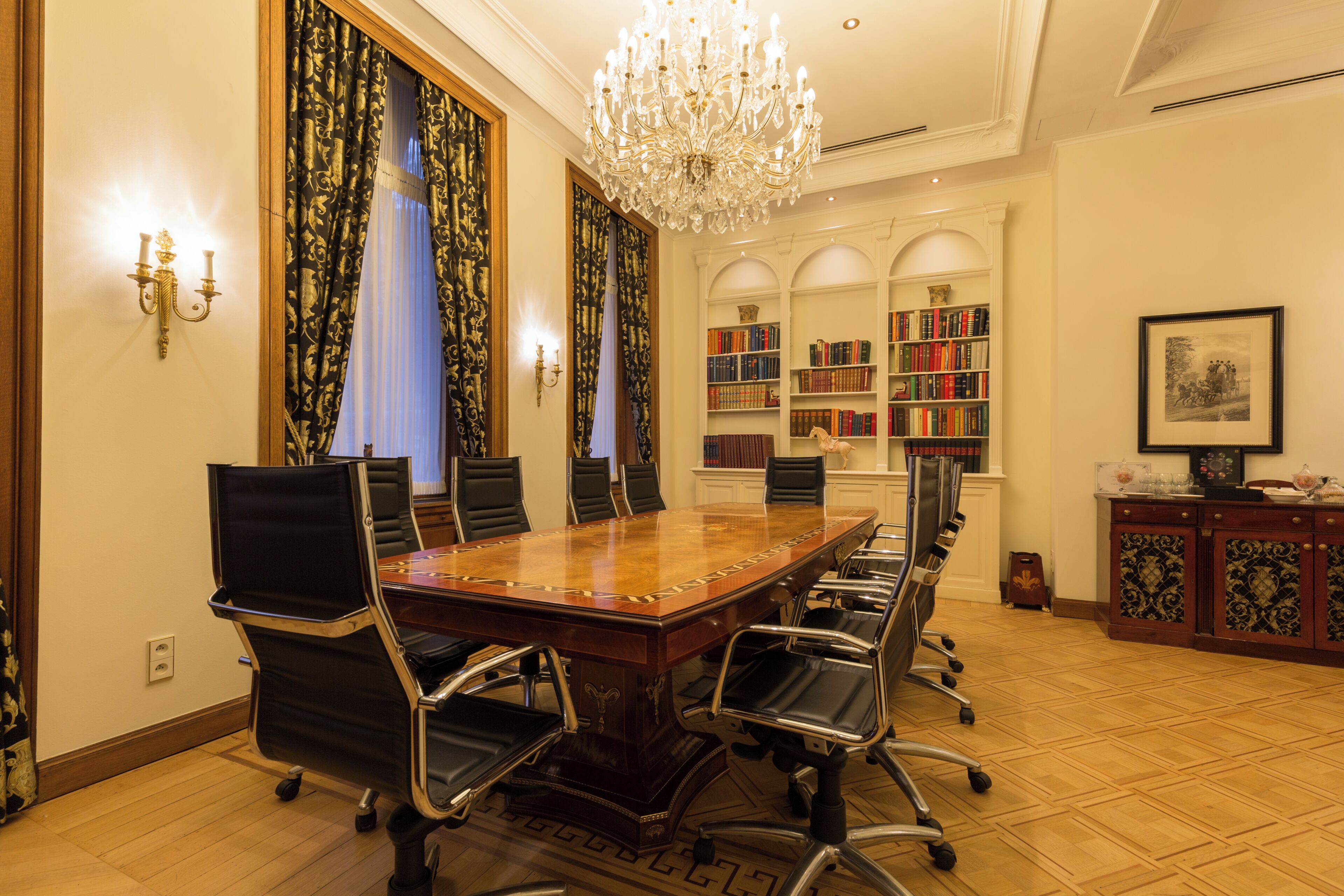 Un salón de conferencias con una gran mesa de madera, sillas negras, una araña y estanterías al fondo.