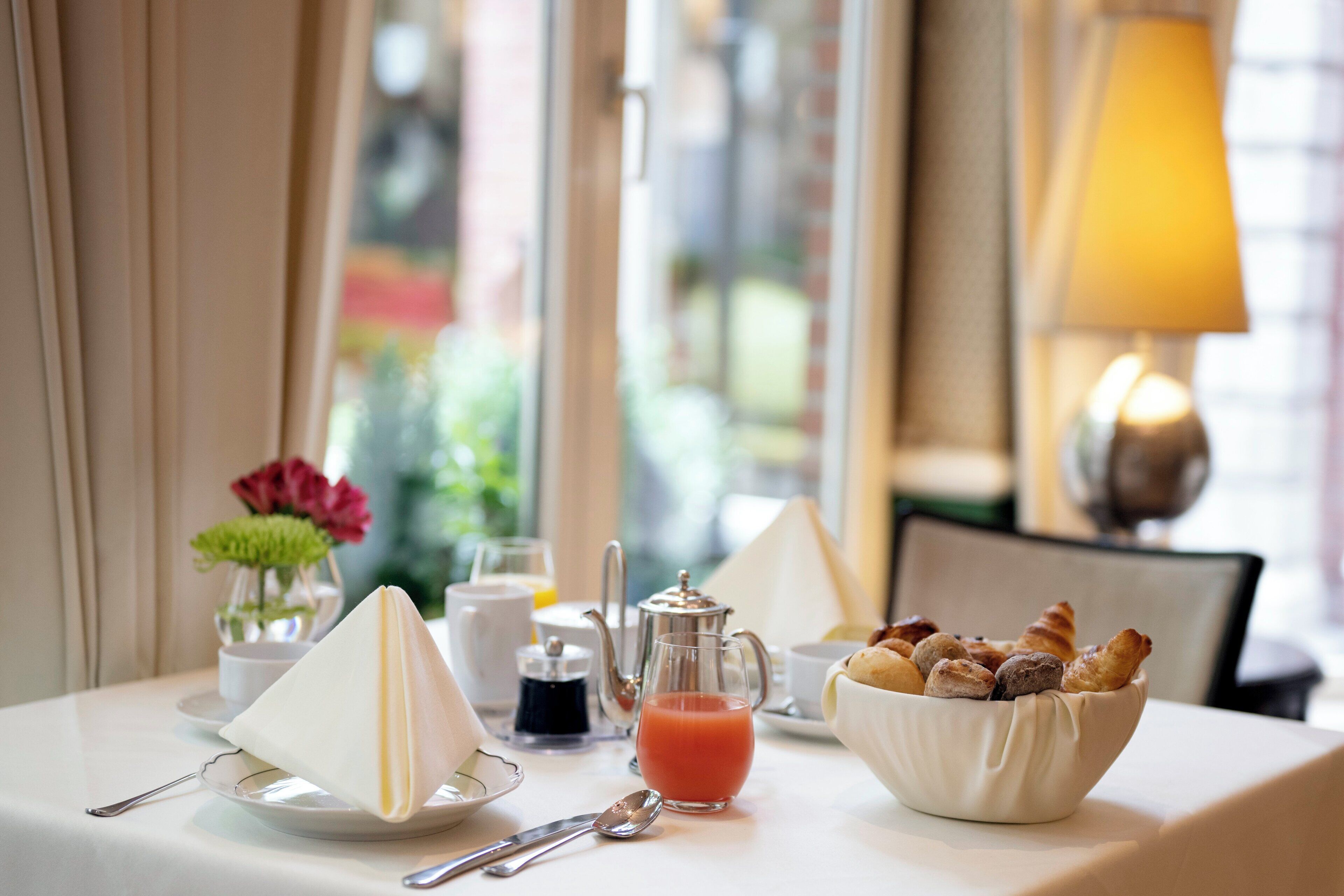 A breakfast table with a teapot, a glass of juice, a bowl of pastries, and a vase of flowers.