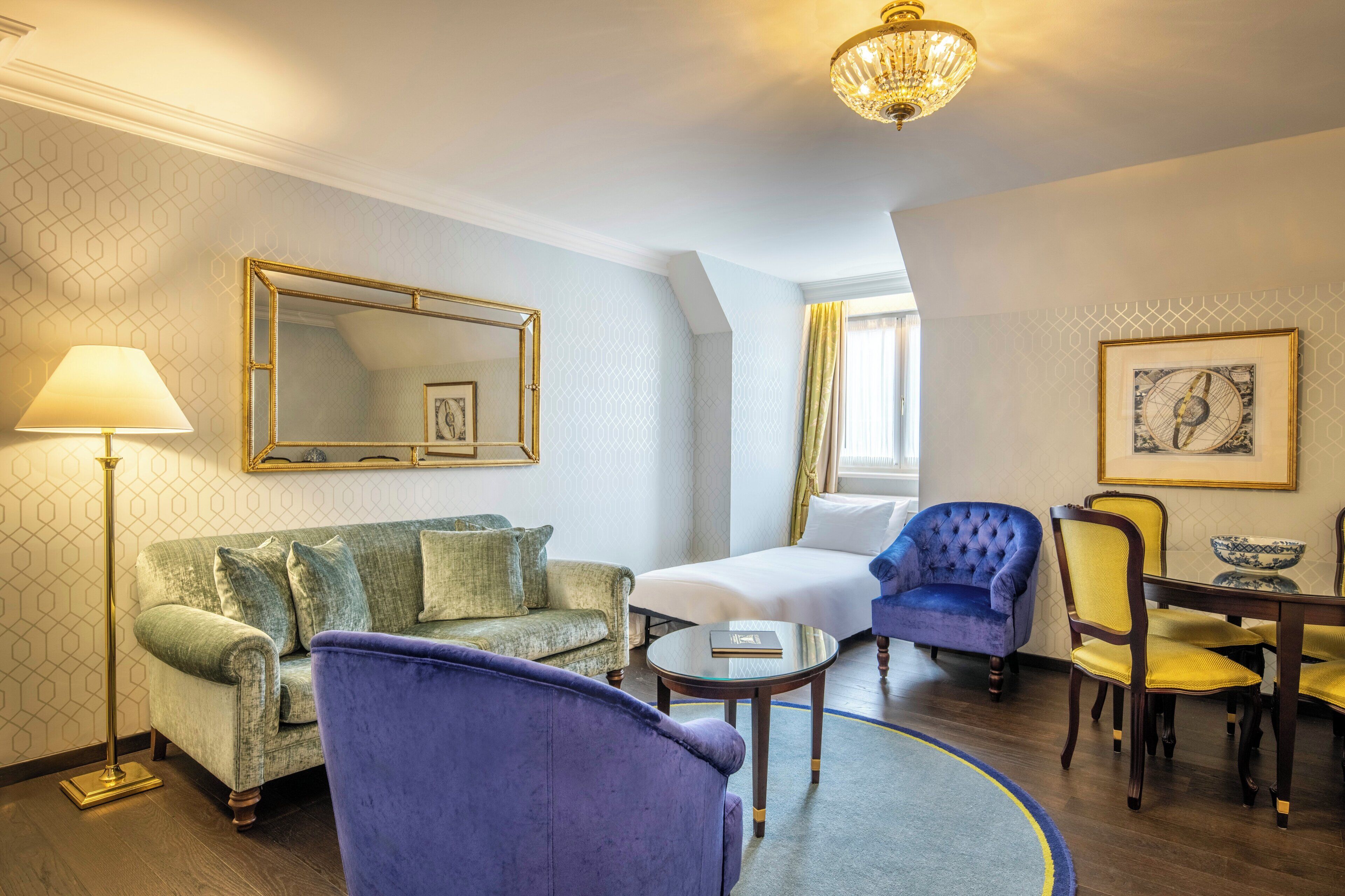 Een hotelkamer met een groene bank, een blauwe fauteuil, een bed, een eettafel met stoelen, een kroonluchter en een grote spiegel aan de muur.
