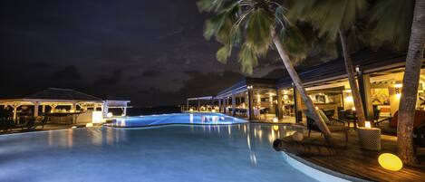 A nighttime view of a resort pool area with a bar, lounge chairs, and palm trees.