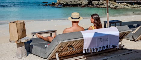 Un couple est assis sur une chaise de plage, une serviette à portée de main, et contemple une mer calme ainsi qu’une côte rocheuse.