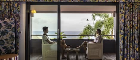 A room with a bed, a balcony with two chairs, and a view of the sea.