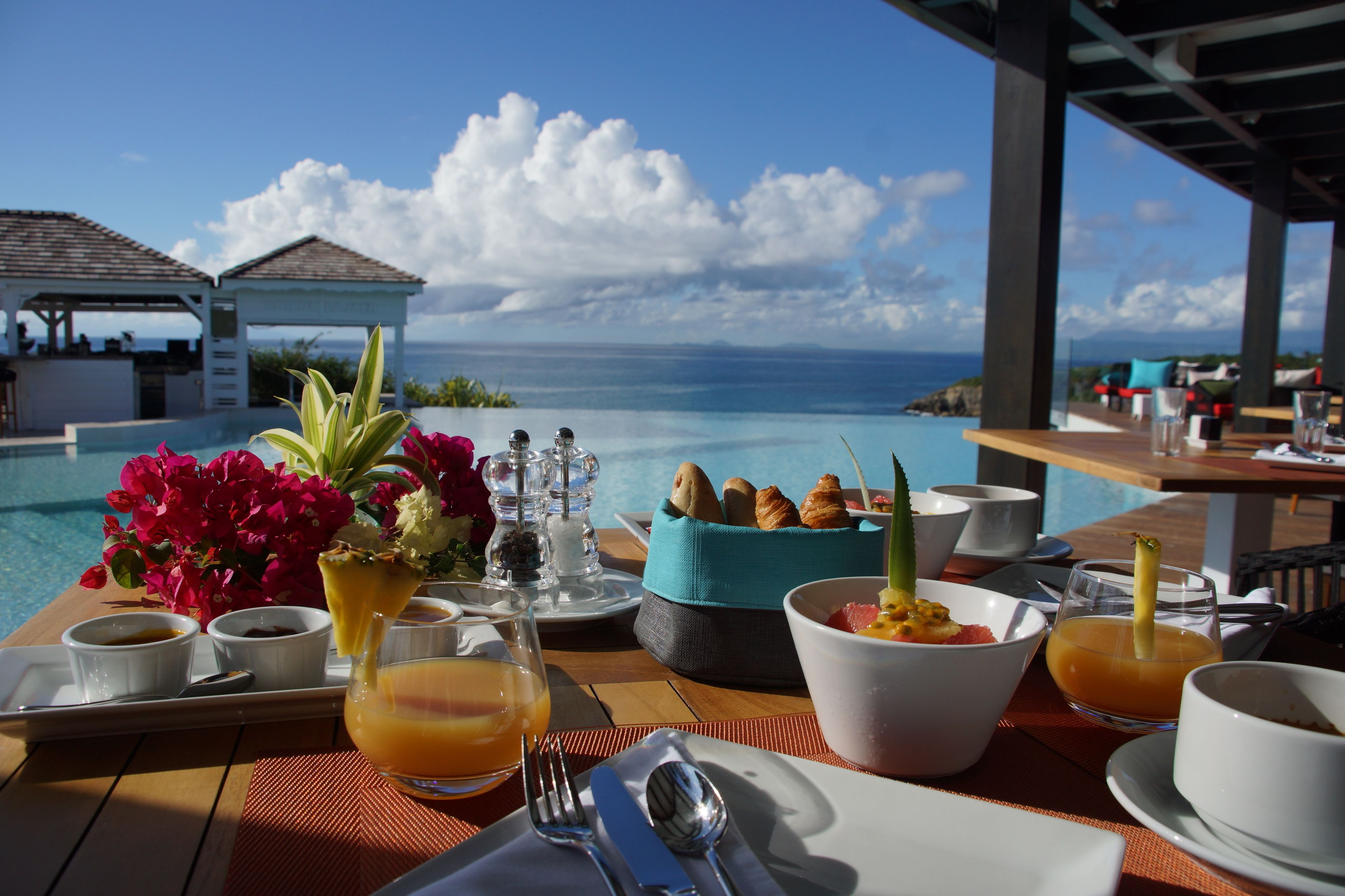 A breakfast setting with a view of the ocean, including a table with breakfast items, a flower arrangement, and a view of the sea.