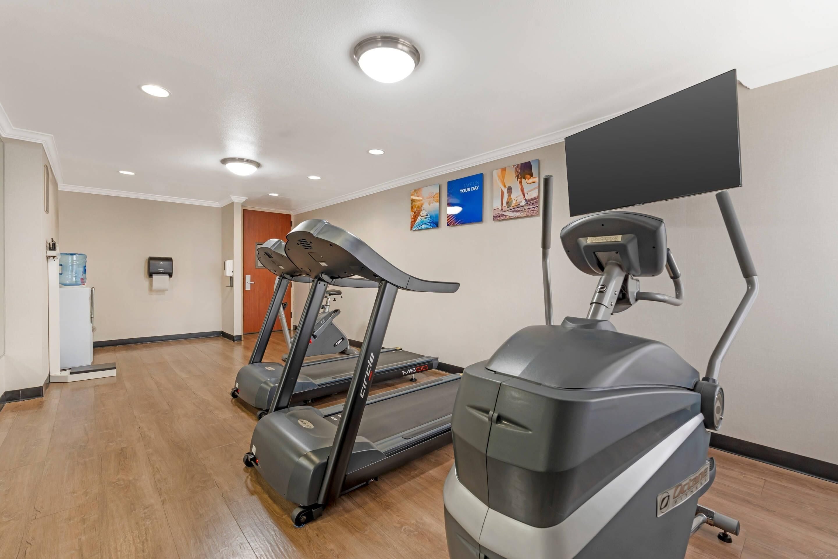 A fitness room with treadmills and a stationary bike, wooden flooring, and a large screen on the wall.