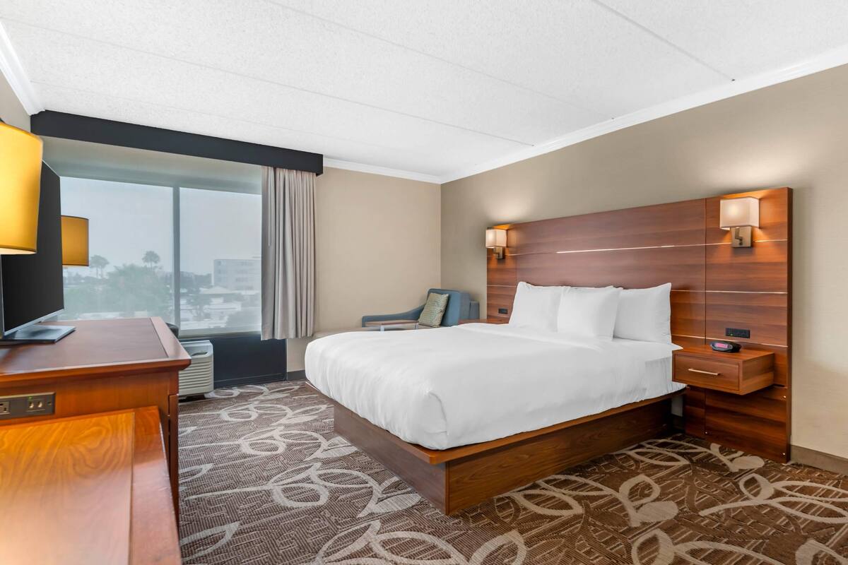 A hotel room with a large bed, a desk, a chair, a nightstand, a lamp, and a view of the city through the window.