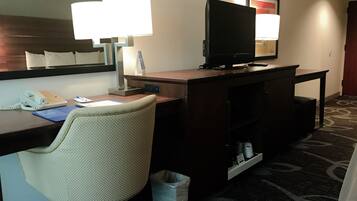 A hotel room with a desk, a chair, a television, a lamp, a phone, and a trash bin.