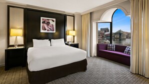 Een hotelkamer met een groot bed, twee bedlampen, een paarse bank en een uitzicht op een stadsbeeld door een rondbogig raam.