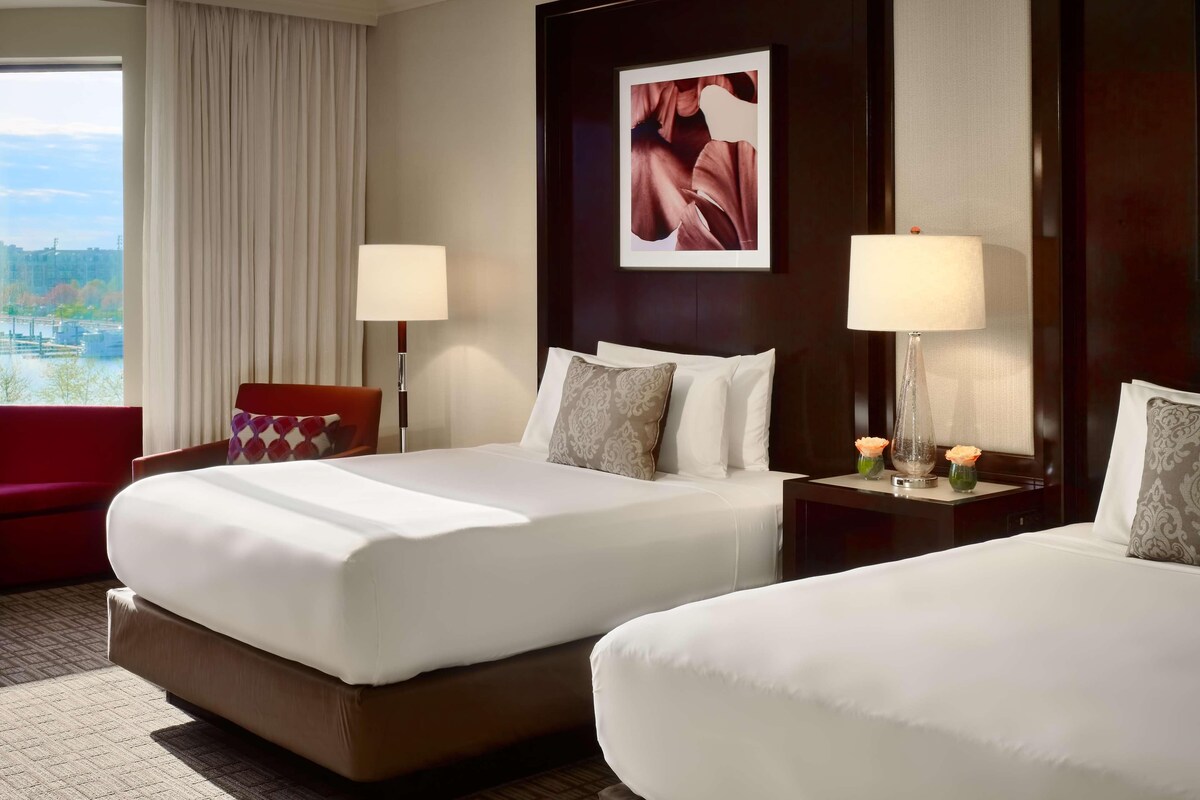 A hotel room with two beds, a large window with a view of a cityscape, a red chair, and a framed picture on the wall.