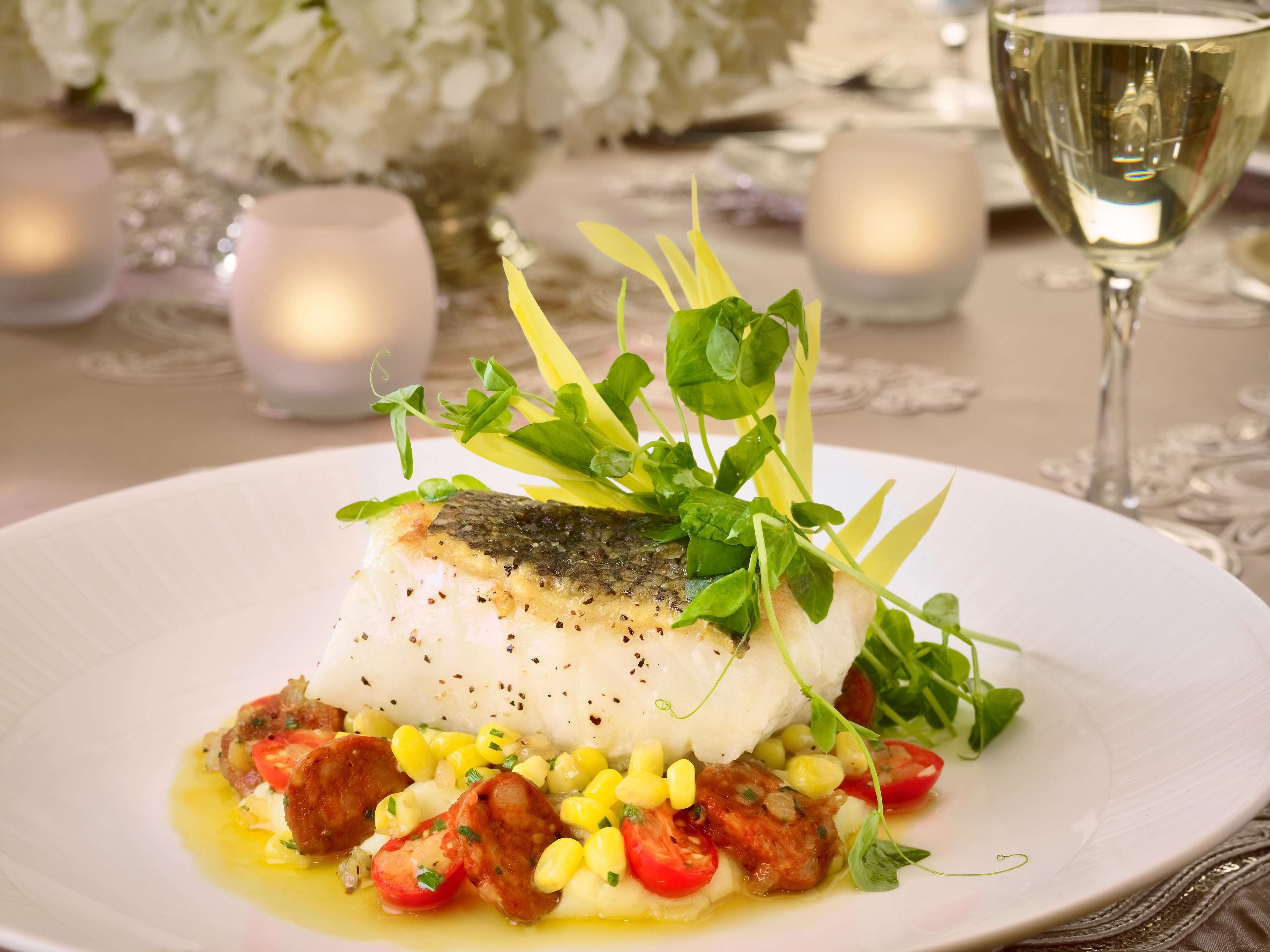 A white plate with a fish fillet, corn, cherry tomatoes, and greens.