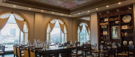 A dining area with tables set for service, large windows with curtains, and a ceiling with detailed artwork.