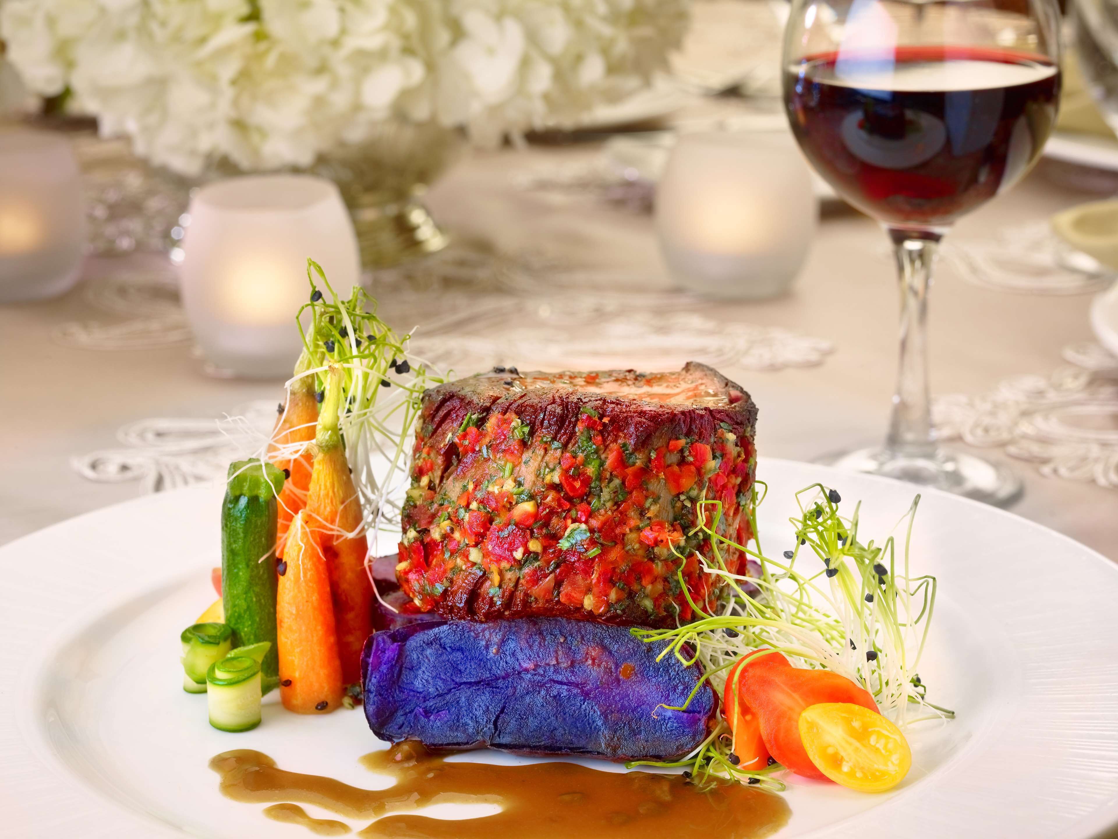 A plated dish with a steak topped with a colorful herb mixture, accompanied by vegetables and a glass of red wine.