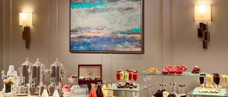 A buffet table with various food items and drink dispensers, a framed abstract painting on the wall, and wall-mounted sconces.