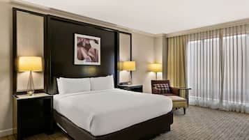 A hotel room with a large bed, two bedside tables with lamps, a chair, and a large window with curtains.