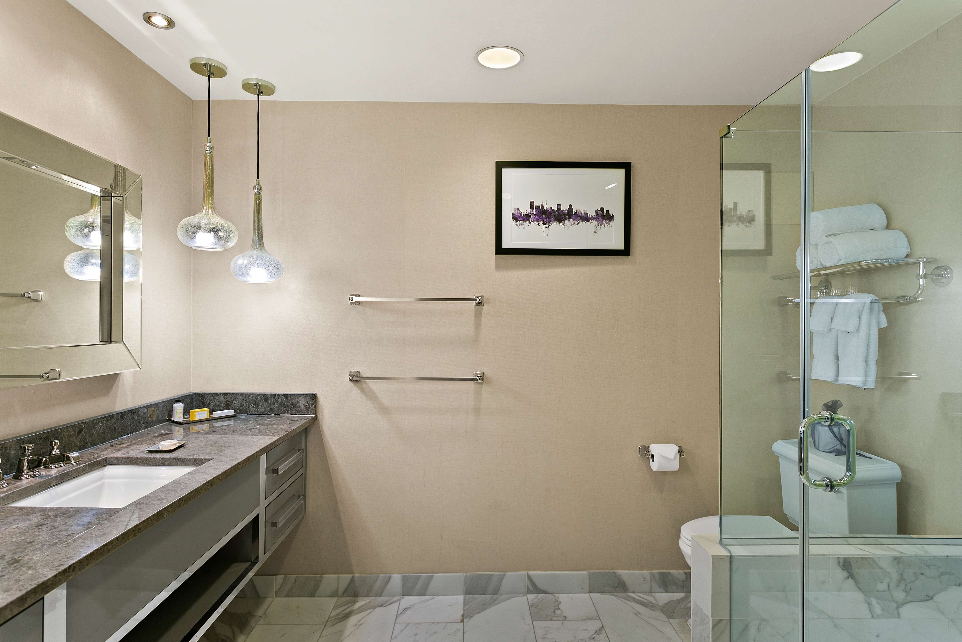A modern bathroom with a large mirror, two sinks, a glass shower enclosure, and a framed cityscape picture on the wall.