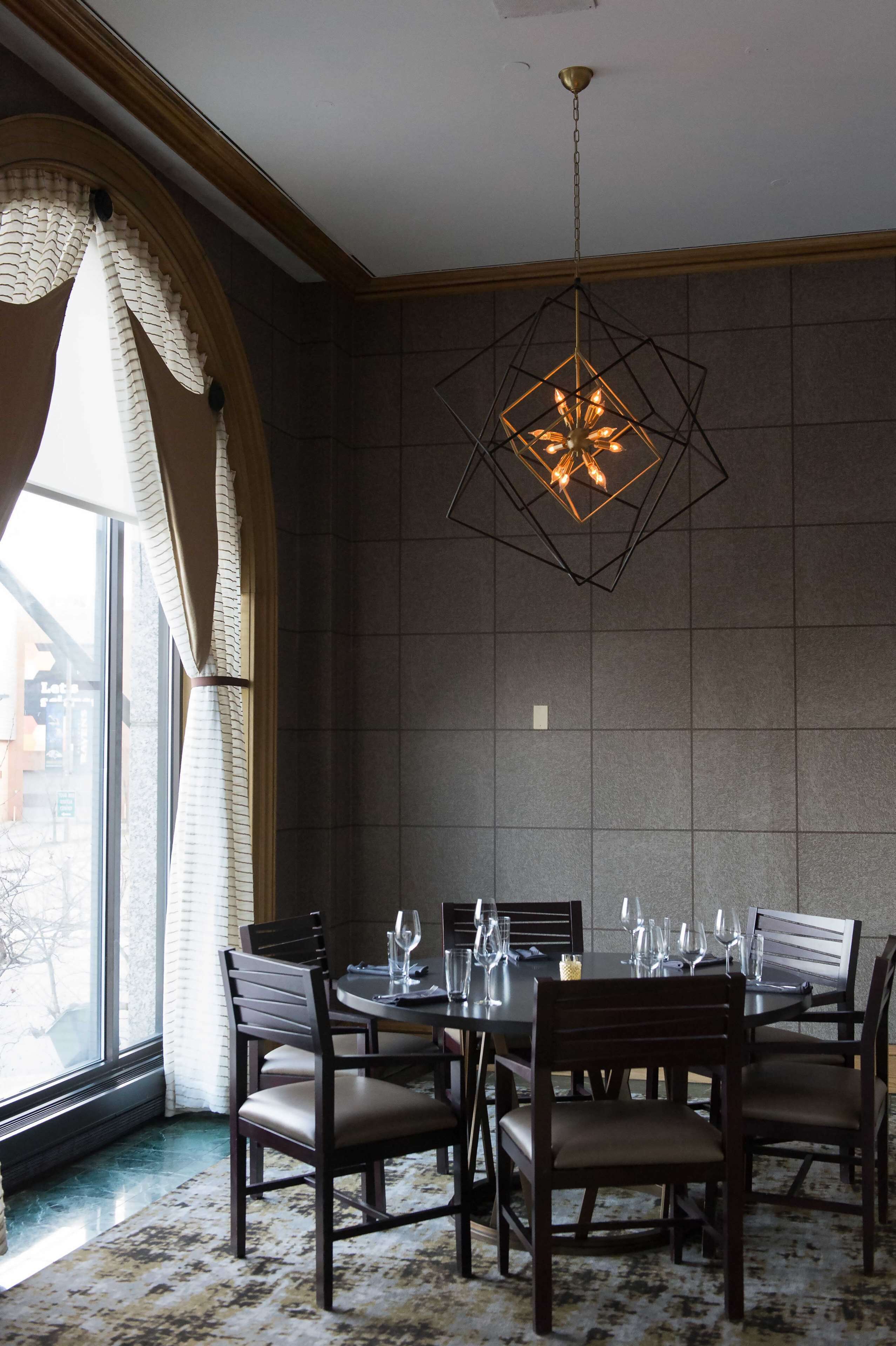 Comedor con una mesa dispuesta para seis comensales, una lámpara colgante de diseño geométrico y vistas a través de una ventana con cortinas.