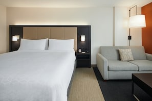 Una habitación de hotel con una cama grande, un sofá gris, una mesita y una mesita de noche con una lámpara.