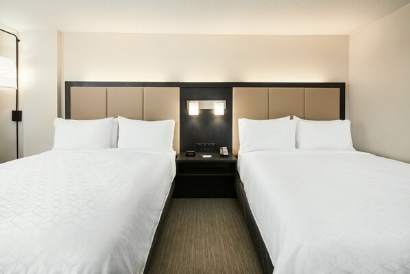 A hotel room with two beds, a nightstand with a phone and lamp, and a headboard with built-in lighting.