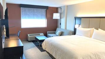 A hotel room with a bed, a desk, a chair, a small table, a heater, and a window with a view.