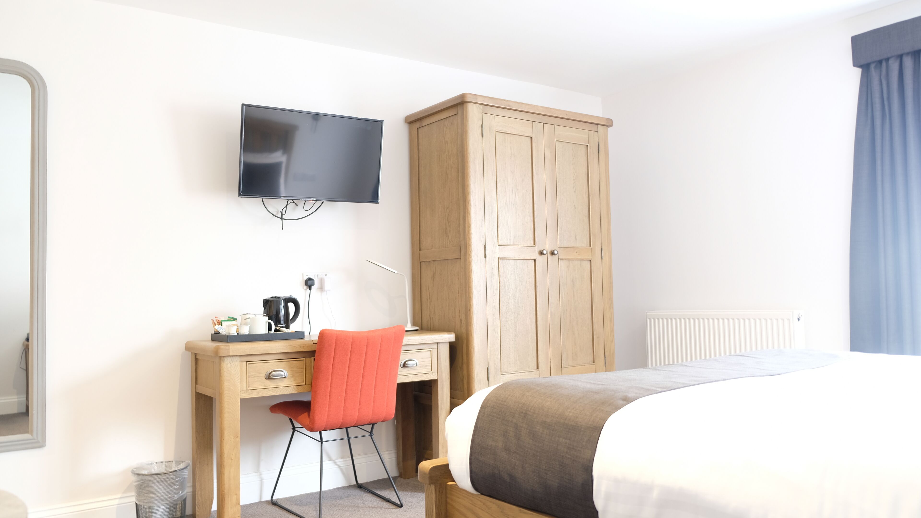 A hotel room with a bed, a desk with a chair, a television, a wardrobe, and a radiator.