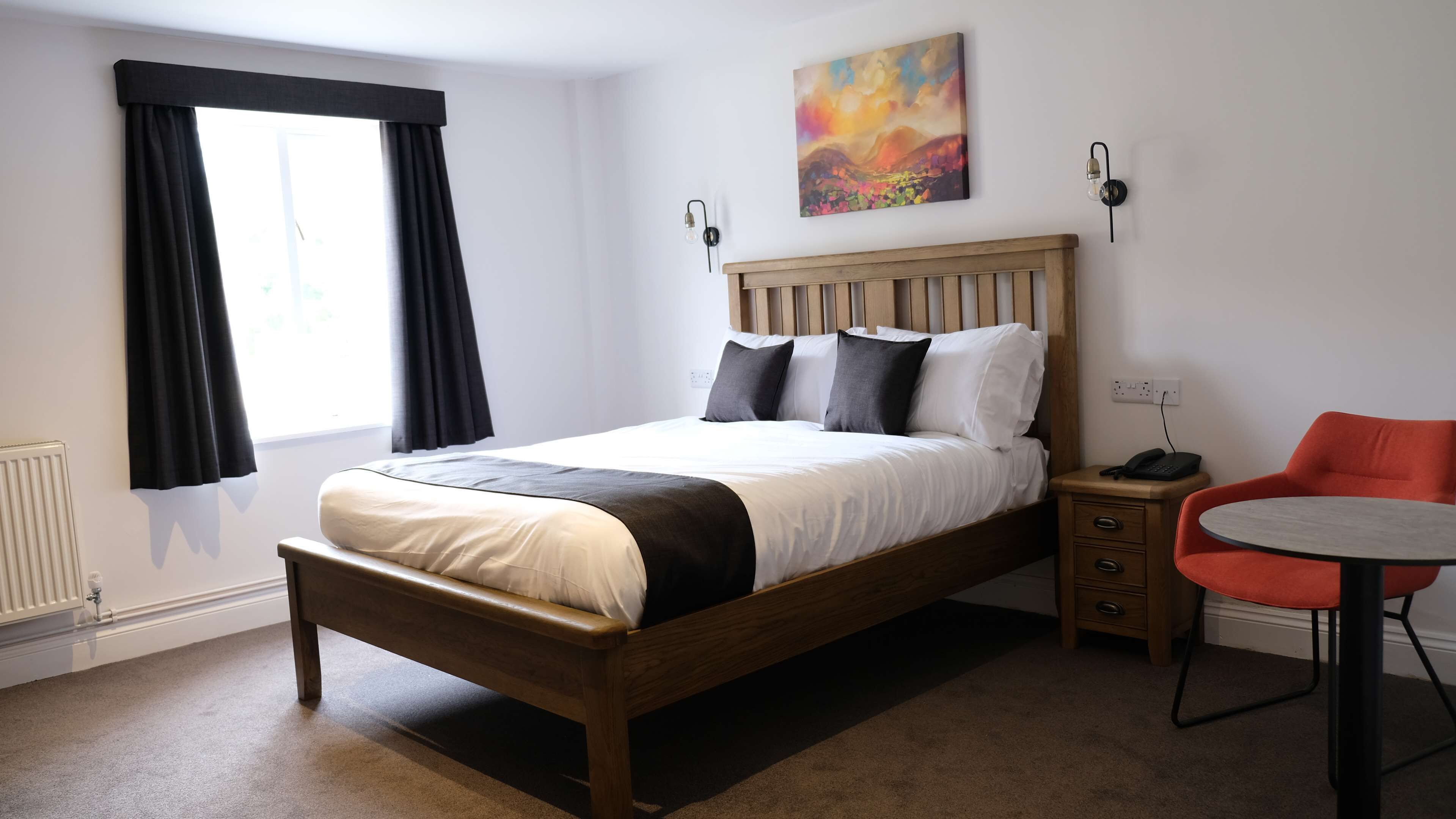 A neatly arranged hotel room with a wooden bed, a small bedside table, a chair, and a round table.