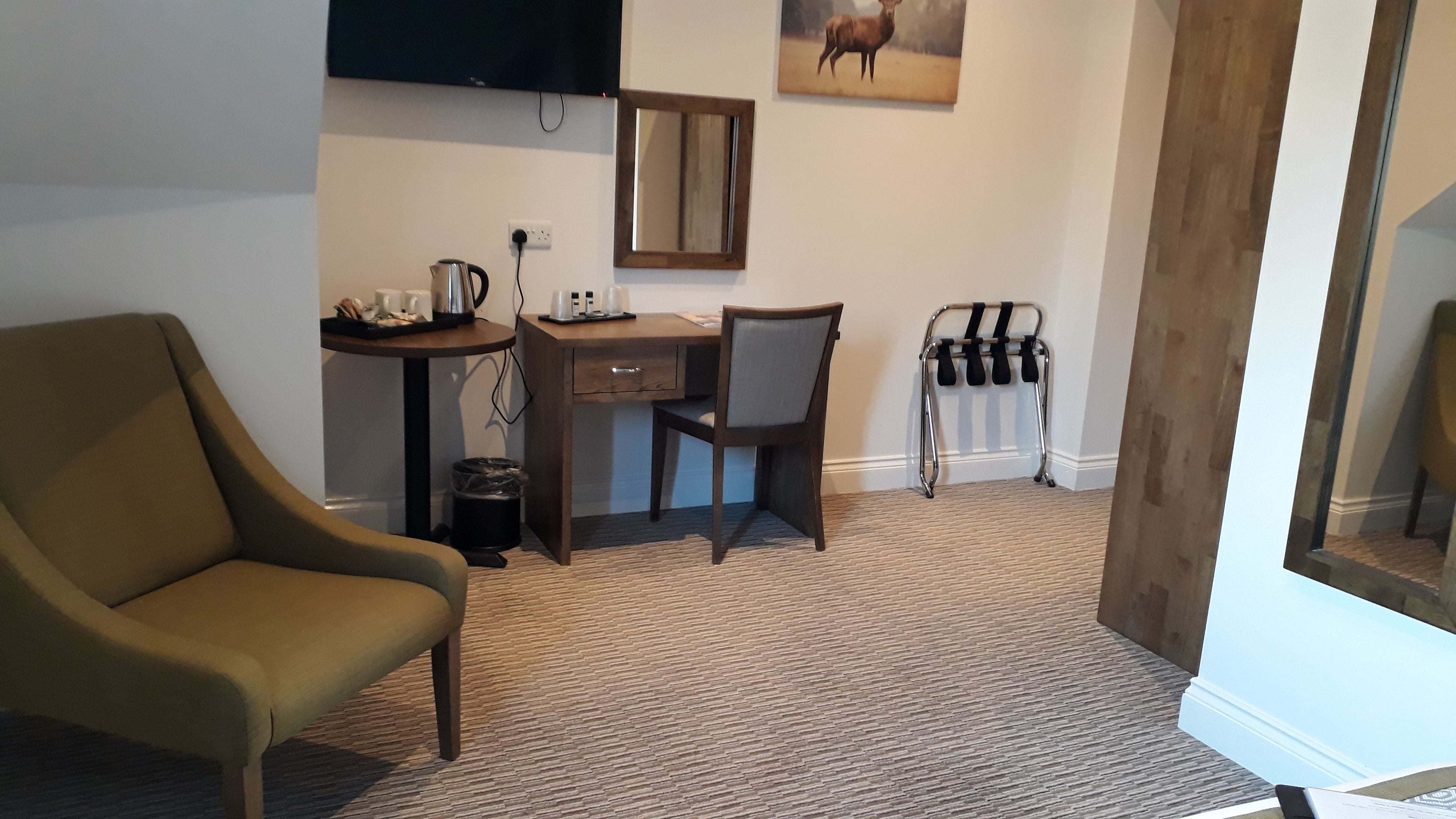 A room with a brown armchair, a wooden desk with a chair, a small table with a kettle and cups, a mirror, and a radiator.