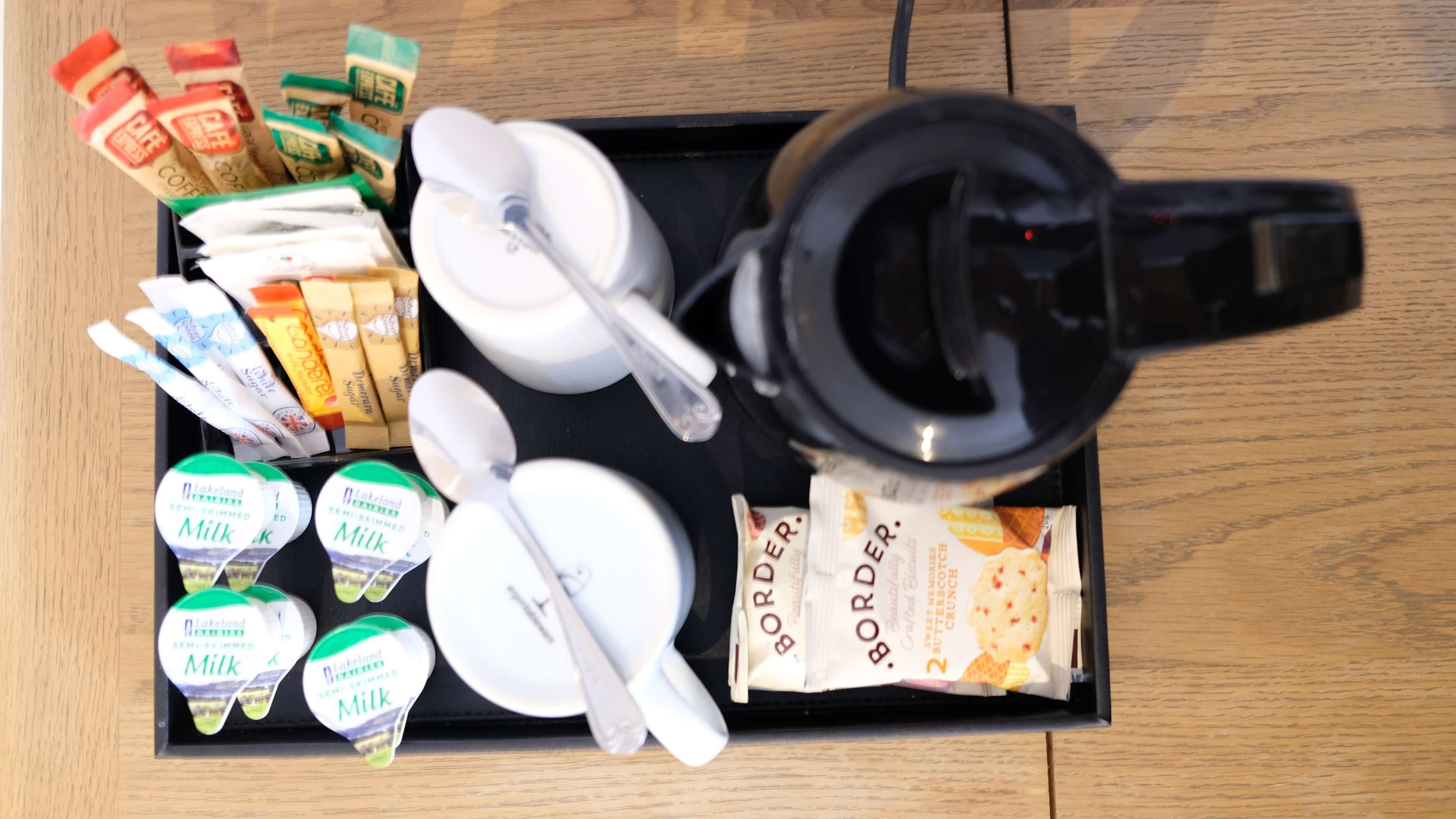 A tray with a coffee maker, sugar packets, instant coffee, a packet of butter, a packet of butter cookies, and containers of semi-skimmed milk.