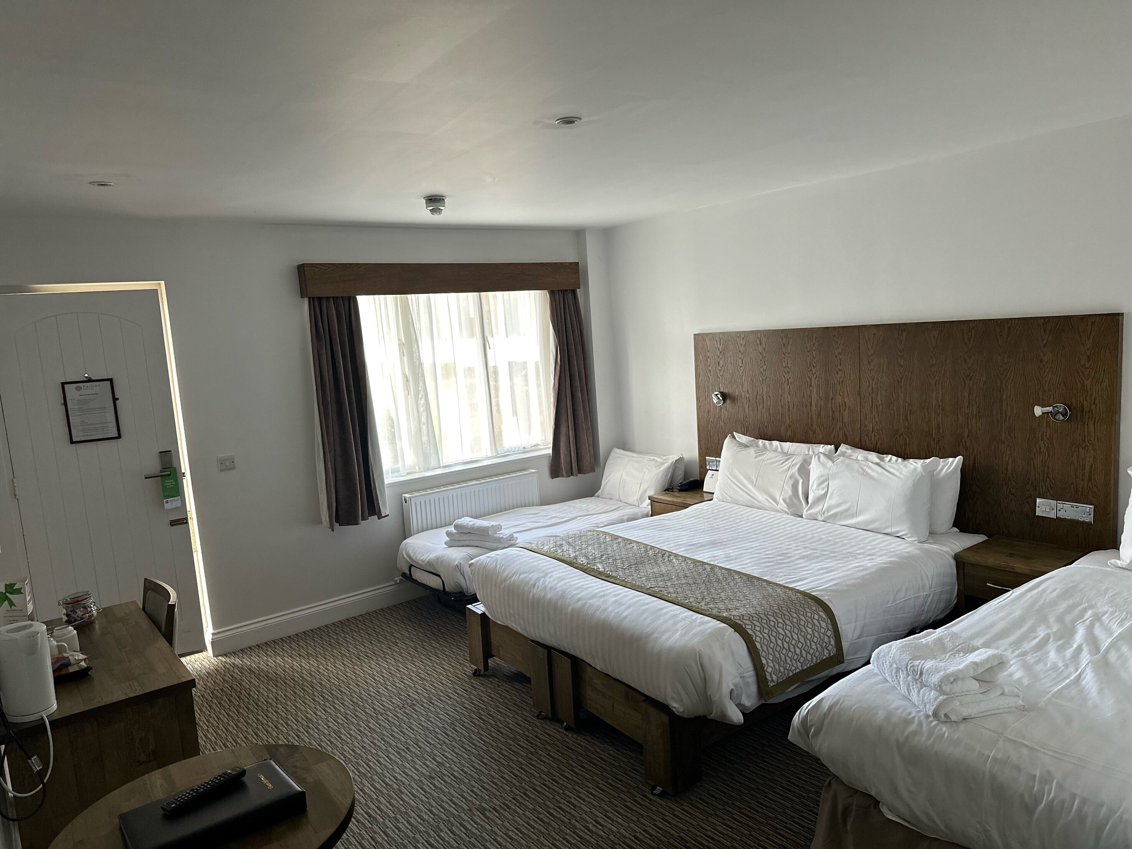 A hotel room with two beds, a wooden headboard, a window with curtains, a bedside table with a lamp, and a desk with a chair and a remote control.
